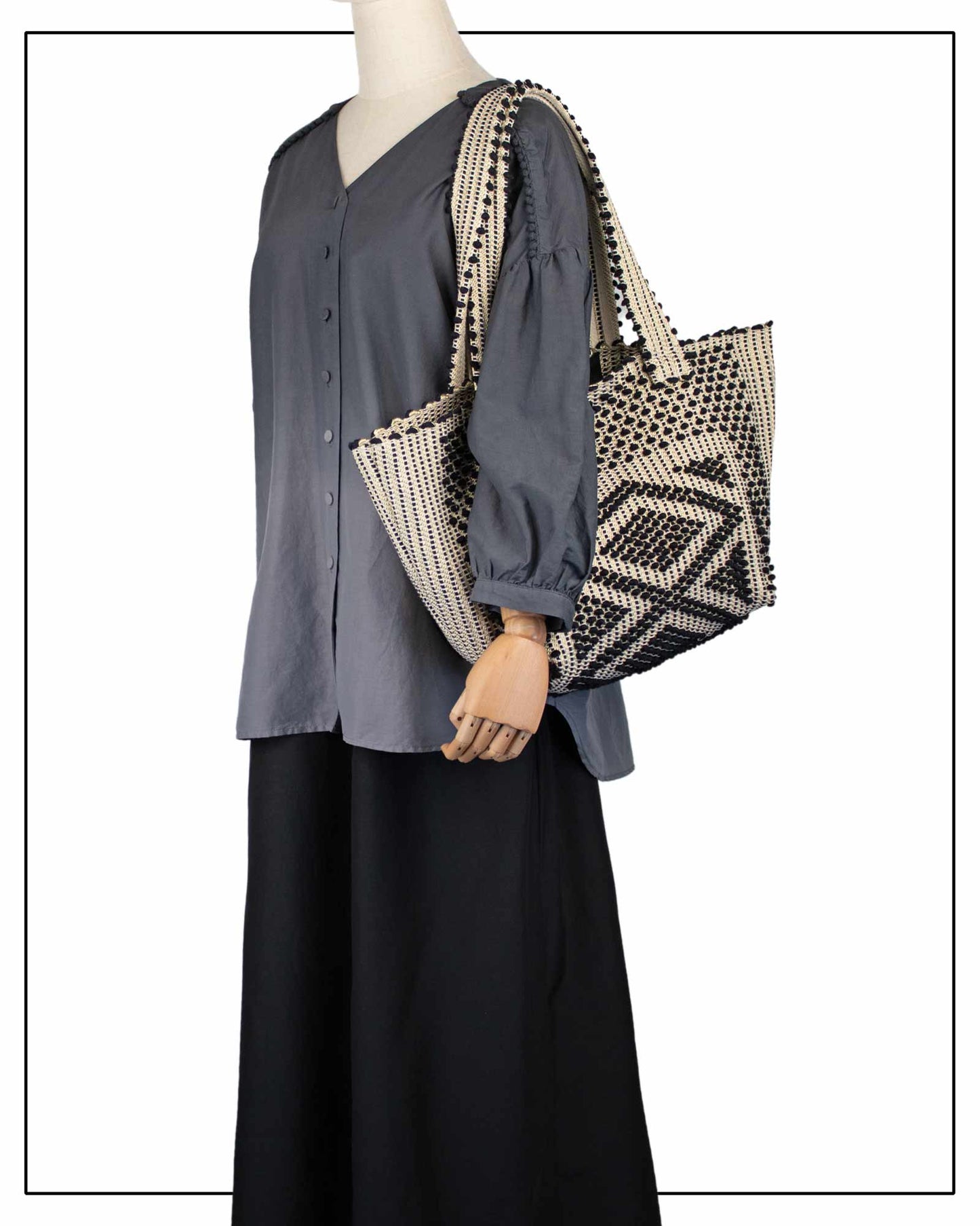 Mannequin wearing a grey shirt and black pants, holding an Antonello Tedde Pula Medium Tote Bag, handwoven in Sardinia with black and golden yarn, a diamond pattern, and a practical flap pocket. 
