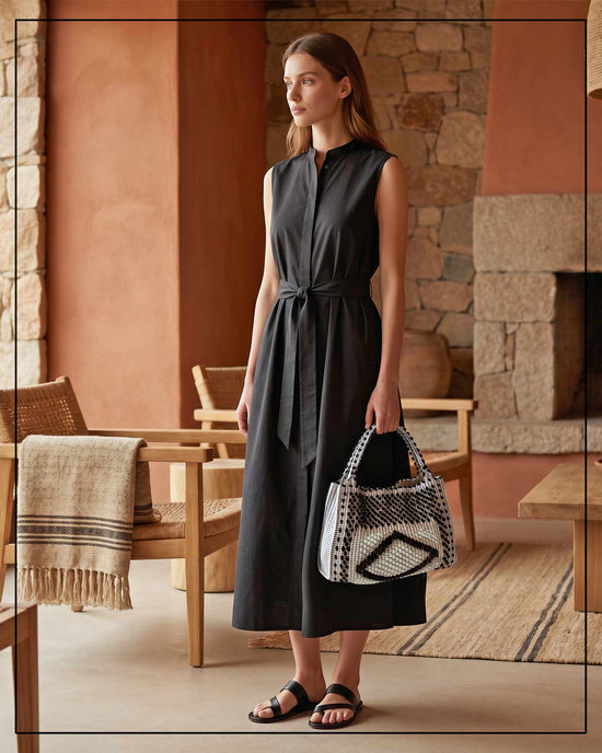Woman in a dark sleeveless dress holding a patterned handbag, all Antonello Tedde, in a rustic interior setting. 