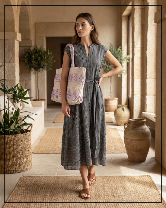 Woman in an Antonello Tedde grey dress holding a patterned tote bag in a sunlit room with plants and stone walls. The handbag is Antonello Tedde Mugoni Zig Zag Tote Bag, handwoven in Sardinia from linen and cotton with geometric zig zag weave and diamond motifs - purple on a cream background. 