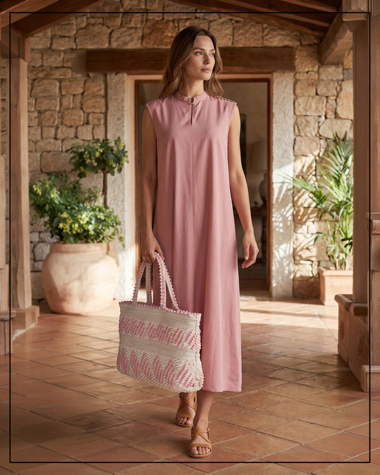 Woman in a pink dress holding a patterned bag in a setting with stone walls and plants. Antonello Tedde Mugoni Zig Zag Tote Bag, handwoven in Sardinia from linen and cotton with geometric zig zag weave and diamond motifs -  frontal view of tote bag with pink on a cream background