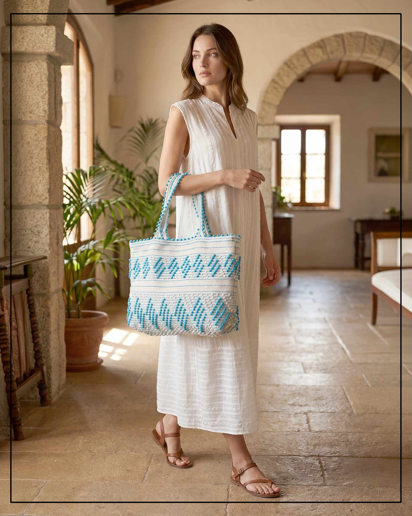 Woman wearing ANTONELLO TEDDE's blue-and-white patterned bag and a cream dress in a sunlit room with stone walls and plants.