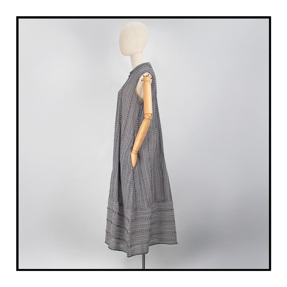 Gray sleeveless dress with a standing collar, crafted from 100% Khadi cotton handwoven in India and made in Italy, featuring a flowing design with pom detail and with hidden pockets and a matching belt for versatile styling.