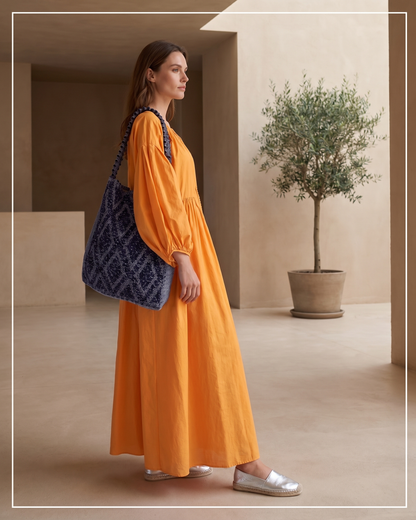 A woman stands indoors wearing a bright orange maxi dress and carrying an Antonello Tedde Bultei handwoven tote bag in black cotton with subtle silver highlights and a textured woven front panel, handcrafted in Sardinia.