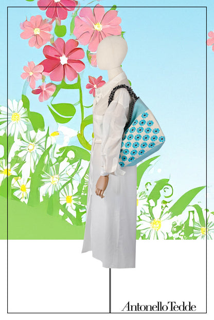 Mannequin wearing a white outfit with a colourful bag against a floral background. view of a turquoise, black and white tote bag with a daisy pattern. Main view of our Sustainable tote - summer bag - luxury handbag - handwoven made in Italy by hand  - timeless individualistic fashion • eco-friendly fashion • socially responsible, lasting fashion.