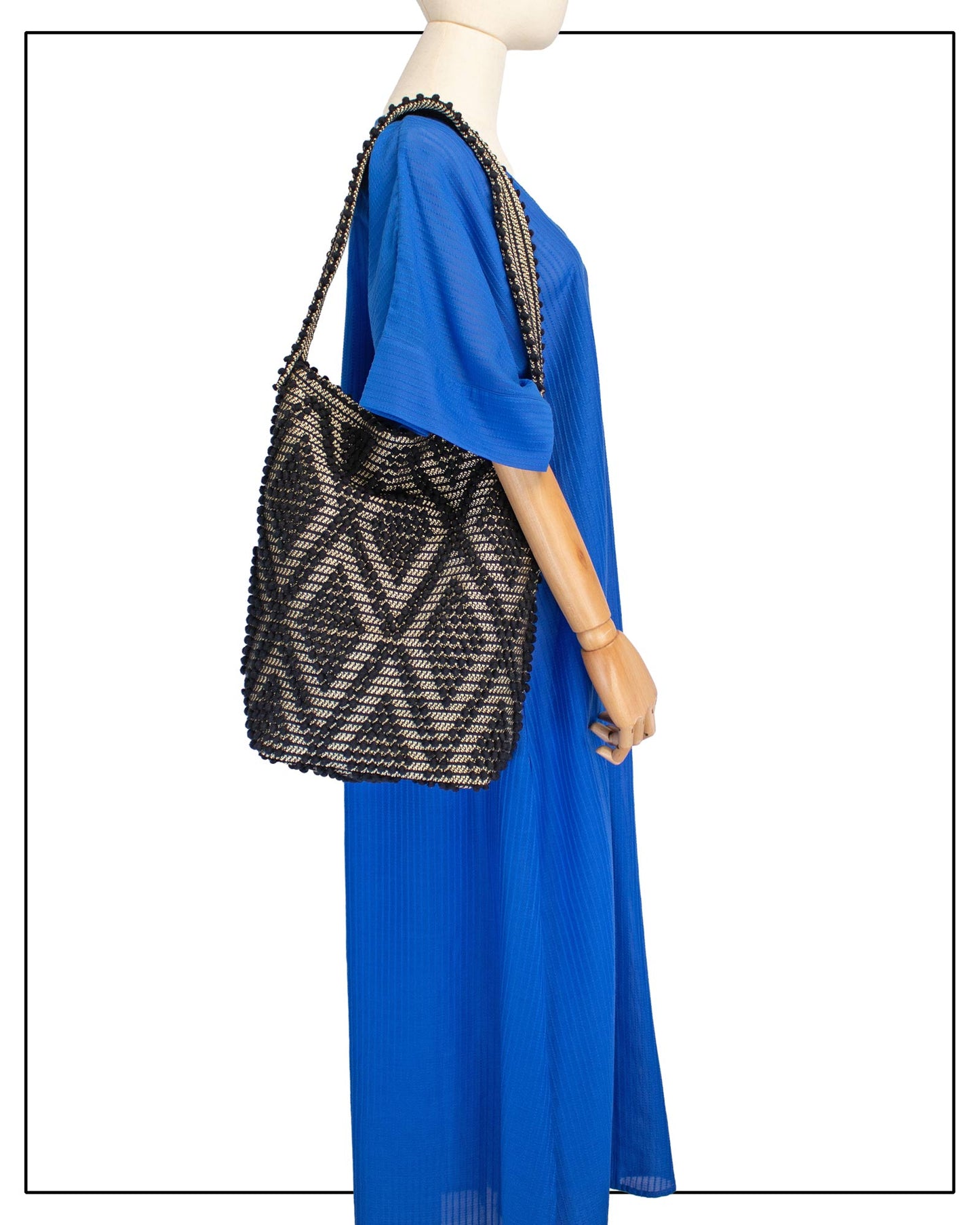 A mannequin wearing a bright royal blue maxi dress with short, wide sleeves and a V-neckline, styled with an Antonello Tedde Bultei Rombi tote bag, handwoven in Sardinia from black cotton with a shimmering golden viscose diamond pattern.