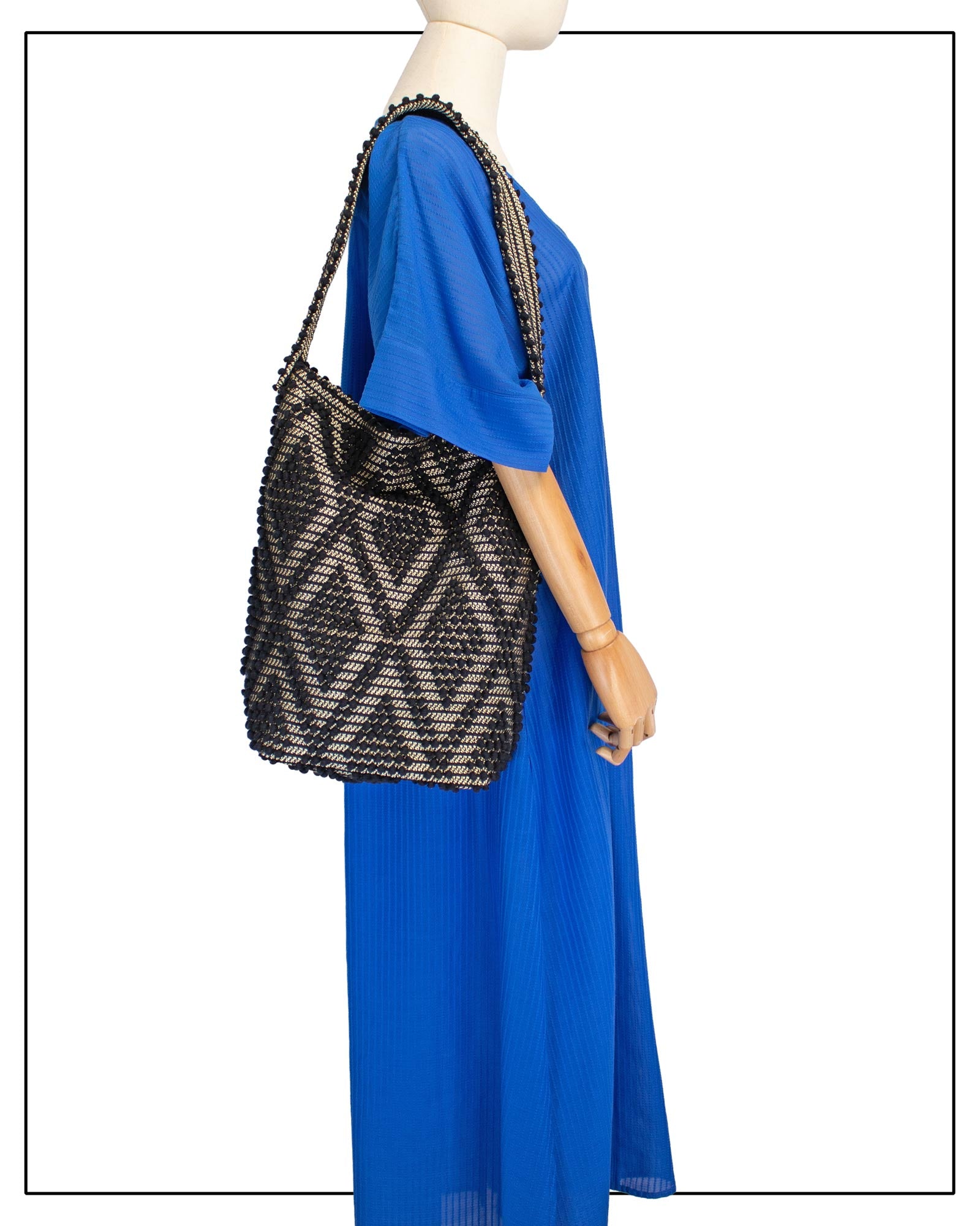 A mannequin wearing a bright royal blue maxi dress with short, wide sleeves and a V-neckline, styled with an Antonello Tedde Bultei Rombi tote bag, handwoven in Sardinia from black cotton with a shimmering golden viscose diamond pattern.