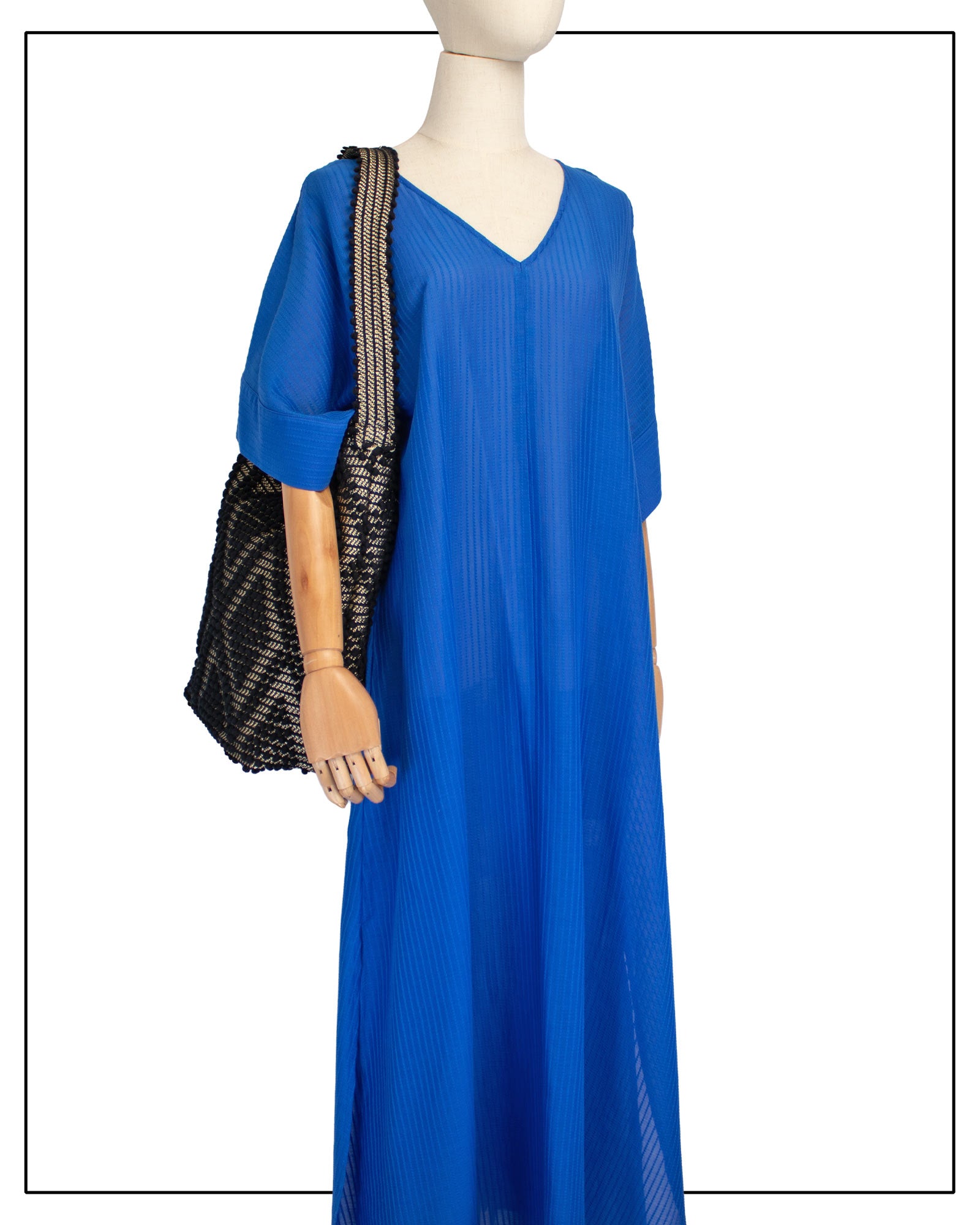 A mannequin wearing a bright royal blue maxi dress with short, wide sleeves and a V-neckline, styled with an Antonello Tedde Bultei Rombi tote bag, handwoven in Sardinia from black cotton with a shimmering golden viscose diamond pattern.