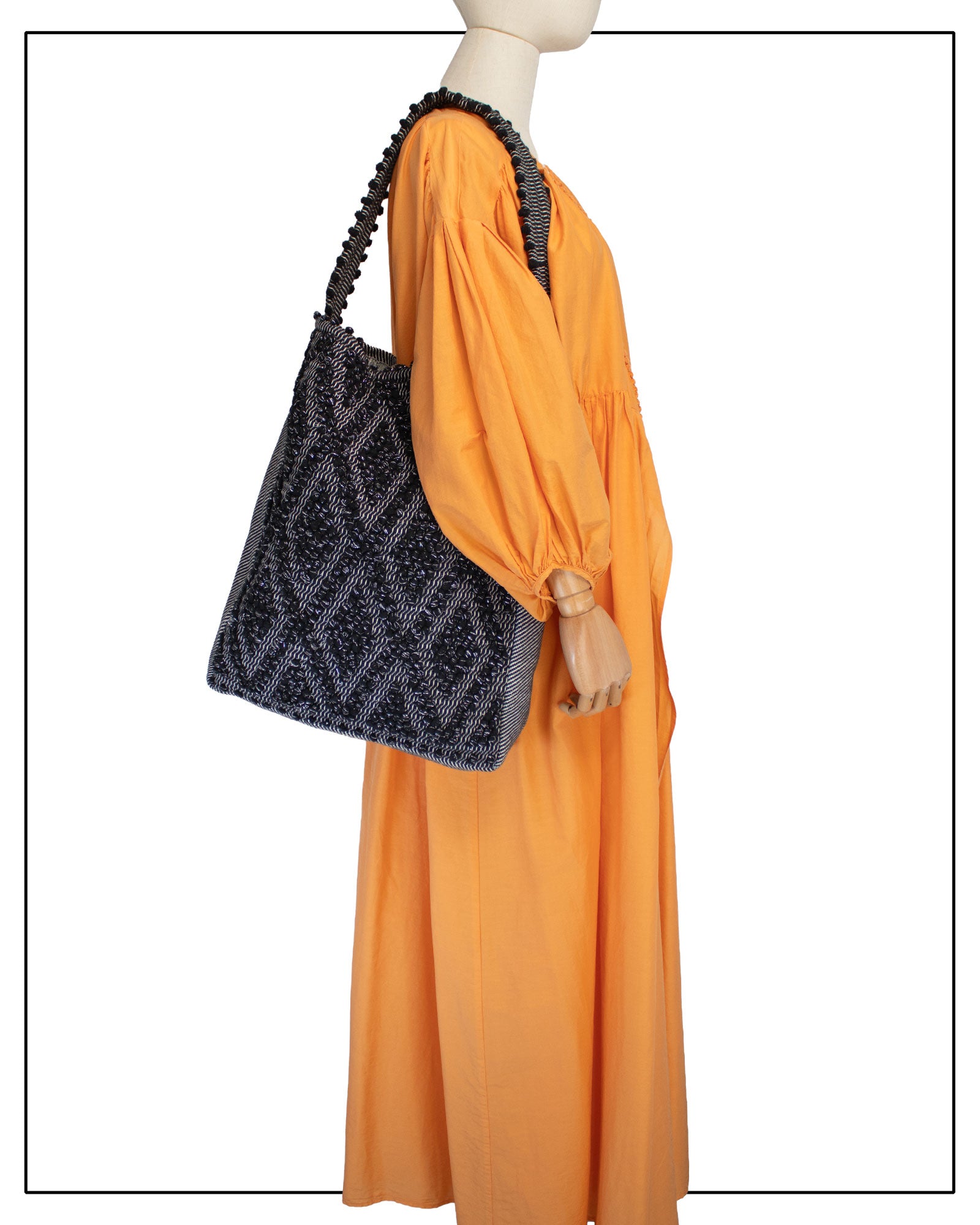 Front view of Antonello Tedde Bultei handwoven tote bag in black cotton with subtle silver highlights and textured woven front panel, handcrafted in Sardinia, shown on a mannequin wearing a long orange dress against a white background.