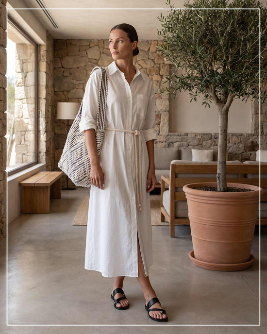A woman stands in a rustic indoor setting wearing a white long-sleeve midi shirt dress with a tie waist belt, carrying an Antonello Tedde Bultei tote bag, handwoven in Sardinia from Italian linen and cotton in cream and blue with timeless artisan craftsmanship.