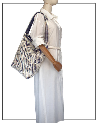 A mannequin wearing a white dress and an Antonello Tedde Bultei tote bag, handwoven in Sardinia from Italian linen and cotton in cream and blue with timeless artisan craftsmanship.