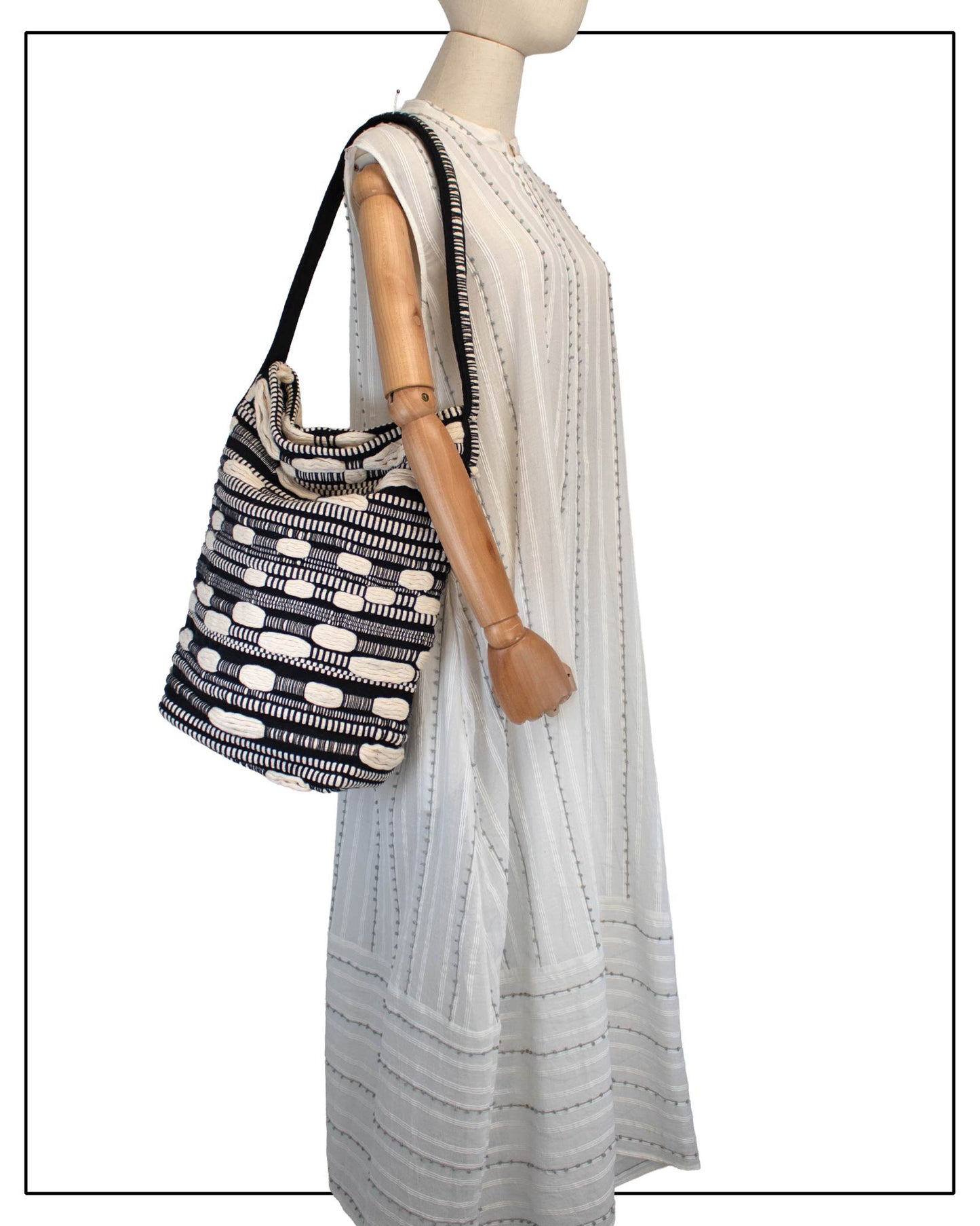 Antonello Tedde Bultei Tote Bag from the Trame in Fuga Collection, handwoven in Sardinia with black and natural cotton, textured woven motifs, and artisanal craftsmanship. Displayed on a mannequin wearing a white embroidered dress against a white background.