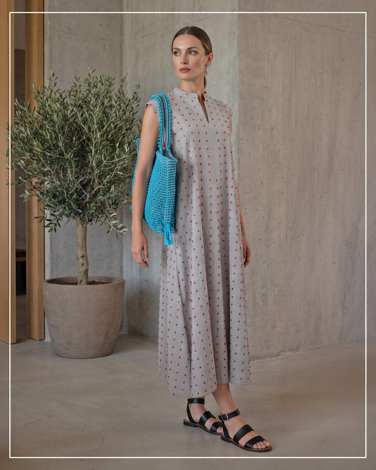A woman stands indoors wearing a sleeveless midi dress with a light gray base and small red and blue polka dots. She is holding a bright blue woven shoulder bag by Antonello Tedde.