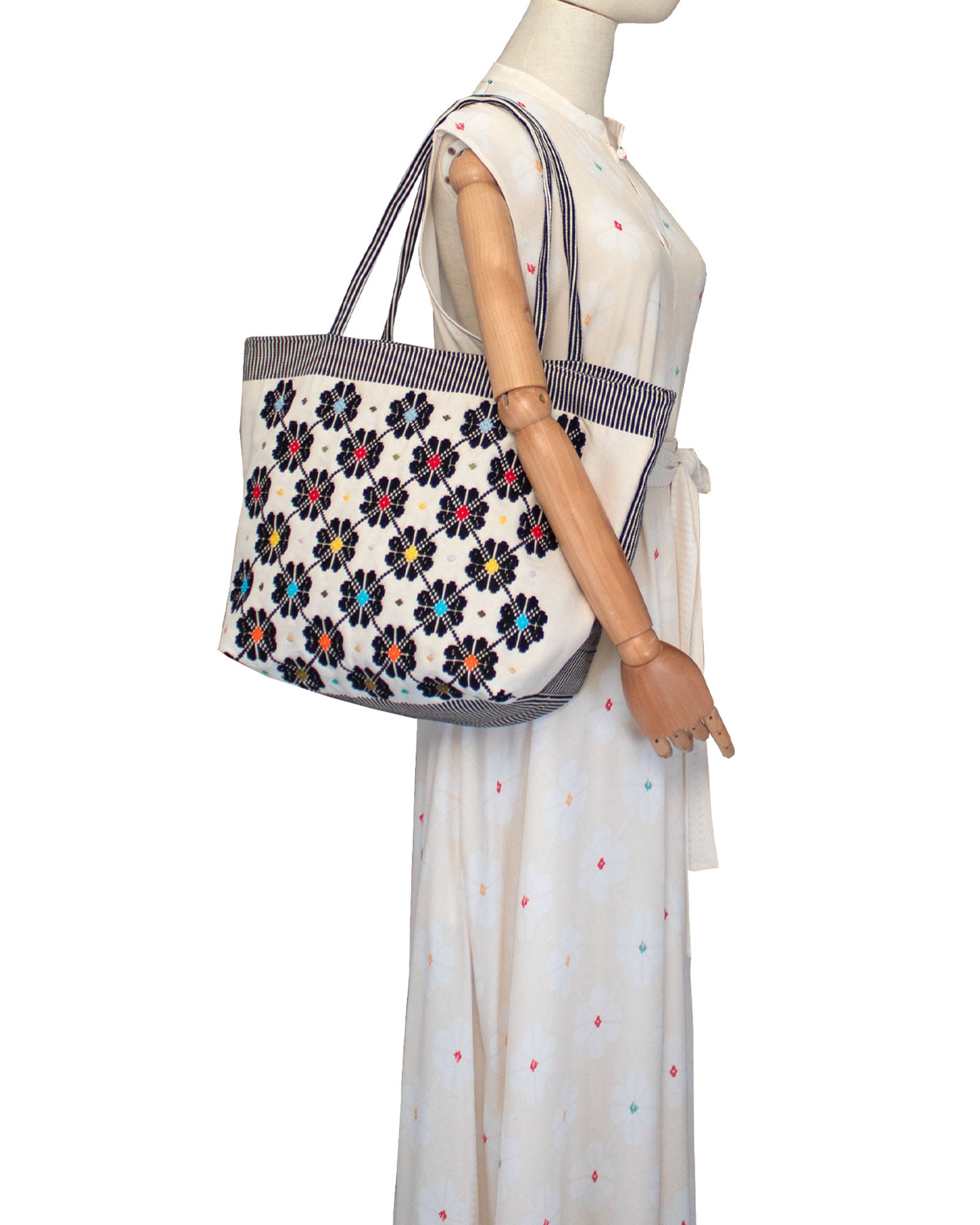 Mannequin wearing a white dress with colourful polka dots holding a floral-patterned tote bag. Front view of a black and white tote bag with a daisy pattern. Main view of our Sustainable tote - summer bag - luxury handbag - handwoven made in Italy by hand  - timeless individualistic fashion • eco-friendly fashion • socially responsible, lasting fashion.
