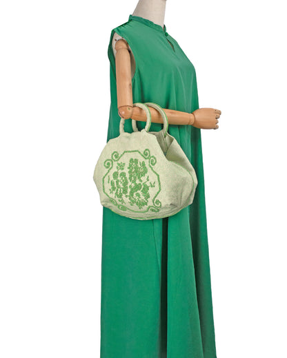 A green handbag with a floral design held by a mannequin against a white background. A handwoven green Faona tote bag with a small pibiones flower bouquet pattern and golden thread was crafted from regenerated cotton by Sardinian artisans. 