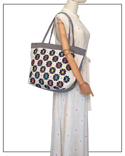 Mannequin wearing a white dress with colourful polka dots holding a floral-patterned tote bag. Front view of a black and white tote bag with a daisy pattern. Main view of our Sustainable tote - summer bag - luxury handbag - handwoven made in Italy by hand  - timeless individualistic fashion • eco-friendly fashion • socially responsible, lasting fashion.
