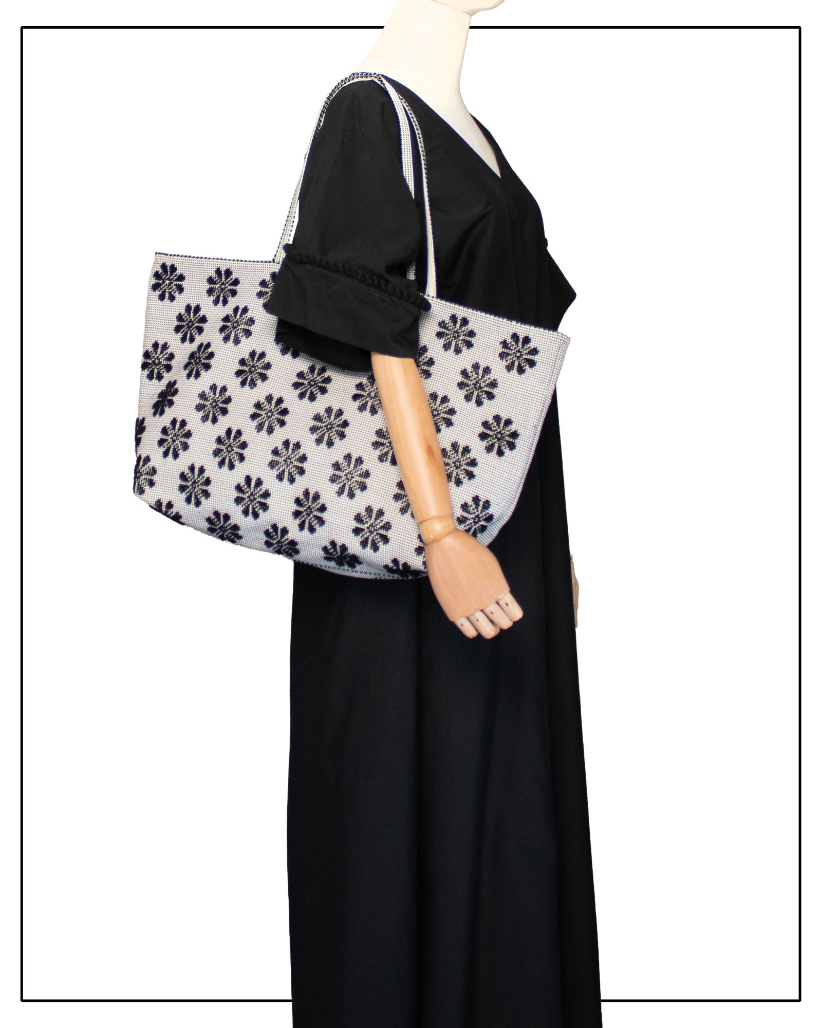 Mannequin wearing a black dress holding a grey tote bag with a floral pattern on a white background. Front view of a black and white tote bag with a daisy pattern. Main view of our Sustainable tote - summer bag - luxury handbag - handwoven made in Italy by hand  - timeless individualistic fashion • eco-friendly fashion • socially responsible, lasting fashion.