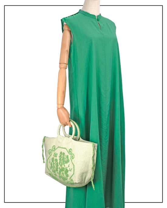 Green dress on a mannequin with a matching green handbag on a white background. A handwoven green Faona tote bag with a small pibiones flower bouquet pattern and golden thread was crafted from regenerated cotton by Sardinian artisans.