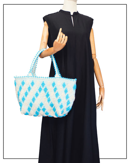 Mannequin wearing a black dress, holding a blue-and-white patterned item. Celvia Medium Torquoise Tote Bag is handwoven, a reimagined classic that marks the return of the beloved rombetti (Multiple Diamonds) pattern in a fresh, contemporary silhouette.