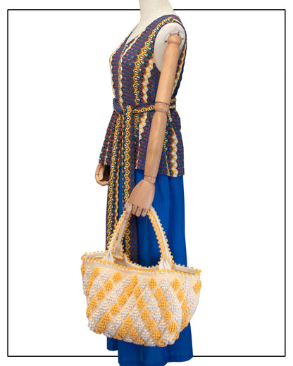 Antonello Tedde's Yellow-and-Cream patterned handbag on a mannequin dressed with Antonello Tedde knit sleeveless top and blue trousers, on a white background. Celvia Medium Red and Cream Tote Bag is handwoven, a reimagined classic that marks the return of the beloved rombetti (Multiple Diamonds) pattern in a fresh, contemporary silhouette.
