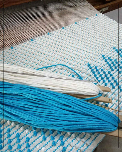 Blue and white yarn on a loom with a woven pattern, where Antonello Tedde Mugoni Zig Zag Tote Bag is handwoven in Sardinia from linen and cotton with geometric zig zag weave and diamond motifs -  frontal view of tote bag with turquoise on a cream background