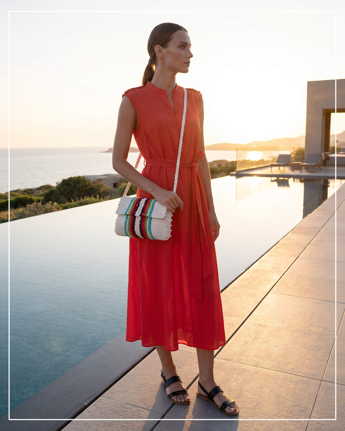A woman stands by an infinity pool at sunset, wearing a sleeveless red midi dress and carrying an Antonello Tedde Suni Arco crossbody bag, handwoven in Sardinia from cream linen and cotton, with colourful raised stripes and a braided shoulder strap.