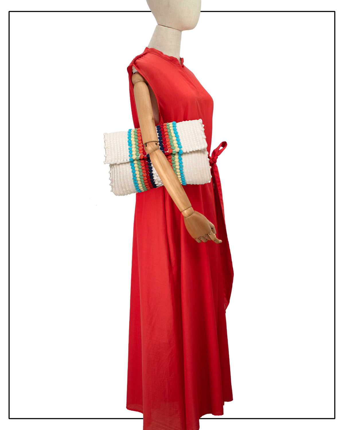 A red sleeveless maxi dress displayed on a mannequin, styled with an Antonello Tedde Suni Arco crossbody bag handwoven in Sardinia in cream linen and cotton with colourful raised stripes and braided shoulder strap.