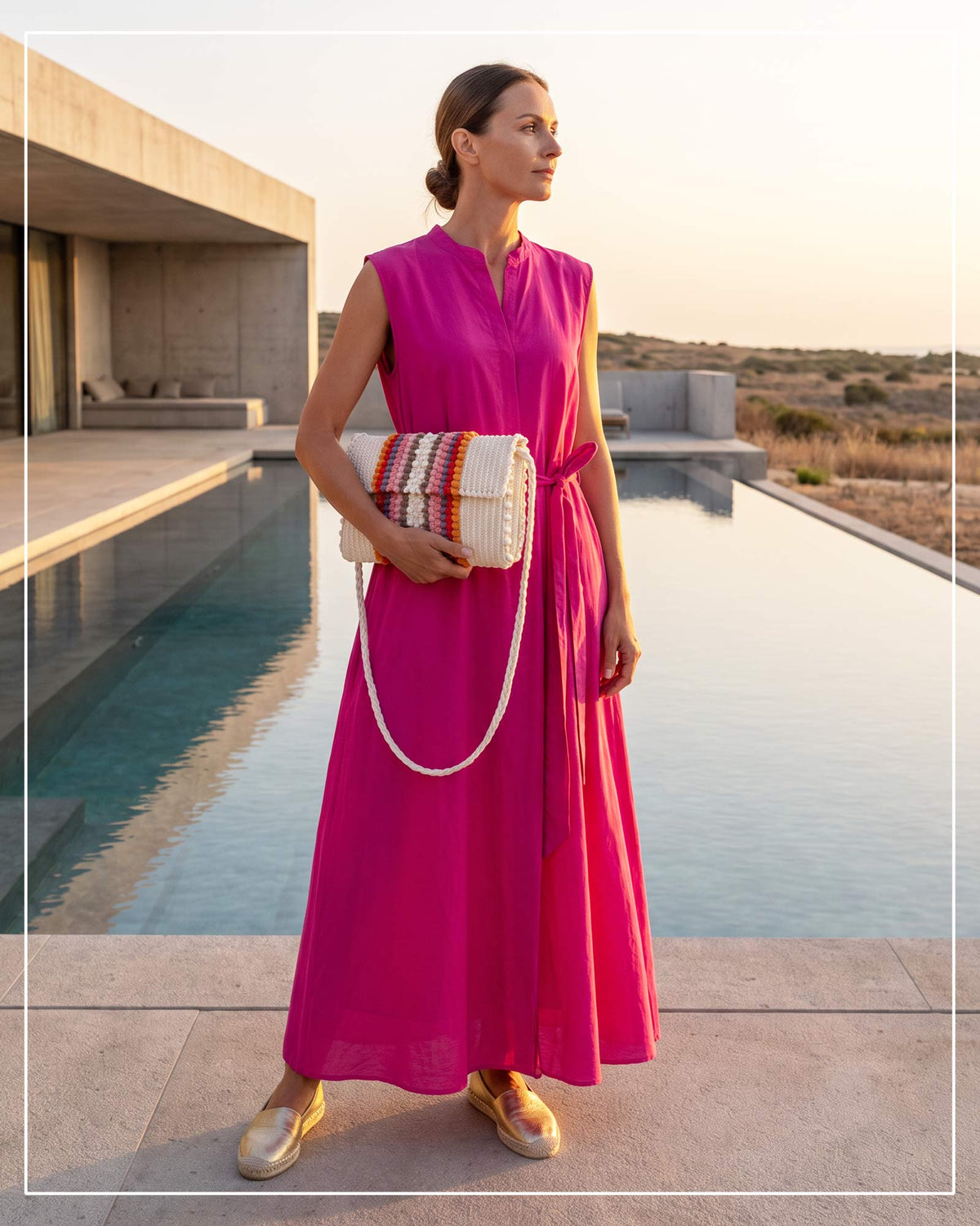 A woman stands by a modern pool at sunset, wearing a sleeveless bright fuchsia maxi dress and a Antonello Tedde Suni Fire crossbody bag handwoven in Sardinia in cream linen and cotton with colourful raised stripes and braided shoulder strap