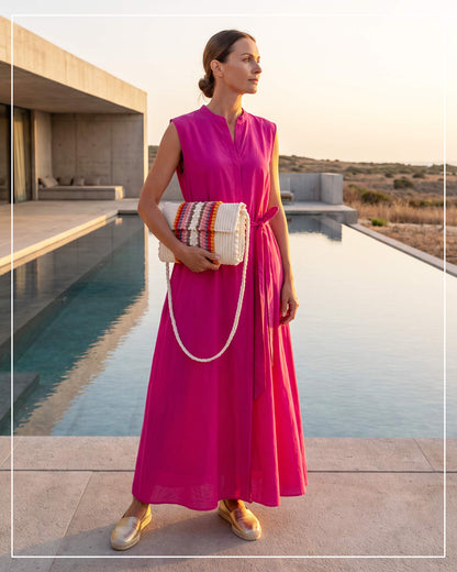 A woman stands by a modern pool at sunset, wearing a sleeveless bright fuchsia maxi dress and a Antonello Tedde Suni Fire crossbody bag handwoven in Sardinia in cream linen and cotton with colourful raised stripes and braided shoulder strap