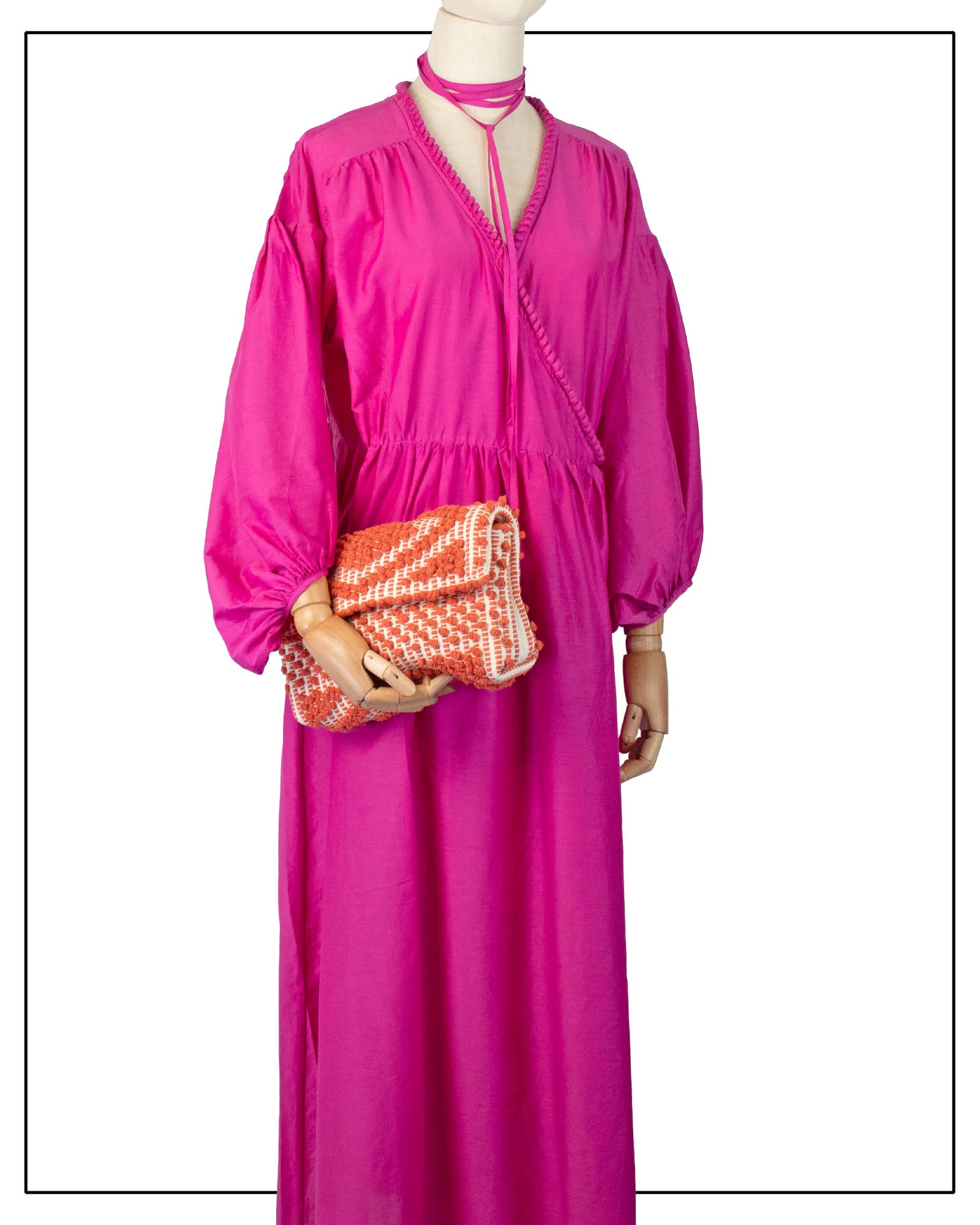 A bright pink maxi dress displayed on a mannequin, styled with a matching pink choker and an Antonello Tedde Suni Square crossbody bag handwoven in Sardinia in coral and cream cotton with a raised geometric pattern and braided strap.