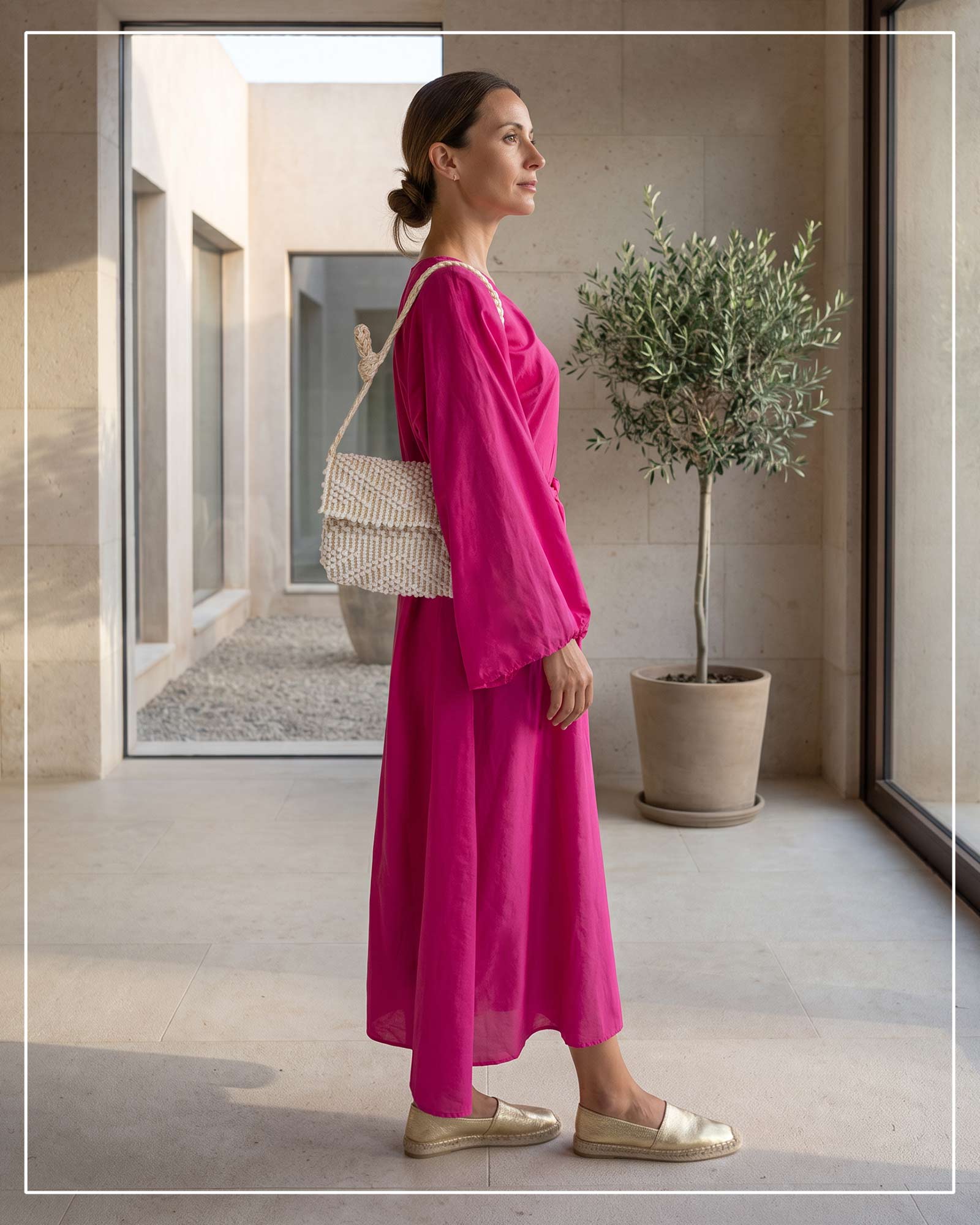 A woman in profile wearing a bright fuchsia two-piece dress with wide sleeves, carrying an Antonello Tedde Suni Small crossbody bag handwoven in Sardinia in cream Italian linen, ivory and subtle golden yarn with braided strap. Wearing gold espadrille flats, standing in a modern architectural corridor with a potted olive tree.