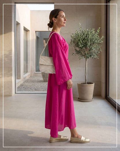 A woman in profile wearing a bright fuchsia two-piece dress with wide sleeves, carrying an Antonello Tedde Suni Small crossbody bag handwoven in Sardinia in cream Italian linen, ivory and subtle golden yarn with braided strap. Wearing gold espadrille flats, standing in a modern architectural corridor with a potted olive tree.