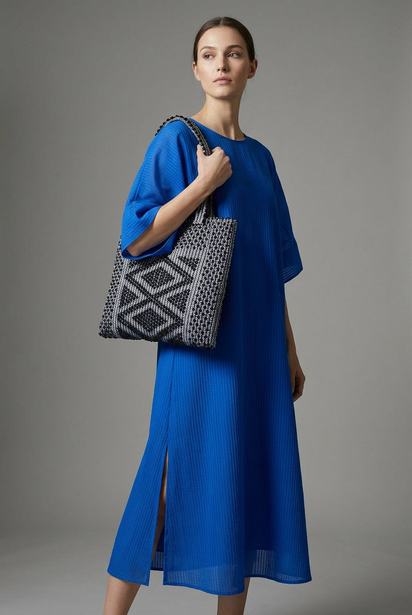 A woman in a blue dress holding a patterned bag against a grey background. Antonello Tedde sculptural handbags in black and silver, combining structured and soft forms, reflecting AW26-AW27 design trends.