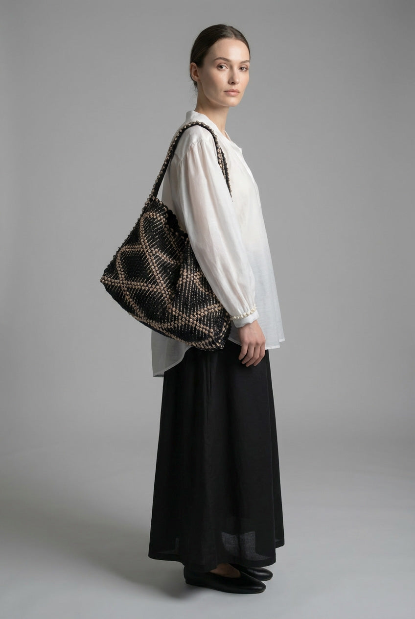 Woman holding a patterned handbag against a plain background. Antonello Tedde handbags in neutral tones, combining structured and soft forms, reflecting AW26 to AW27 design trends.