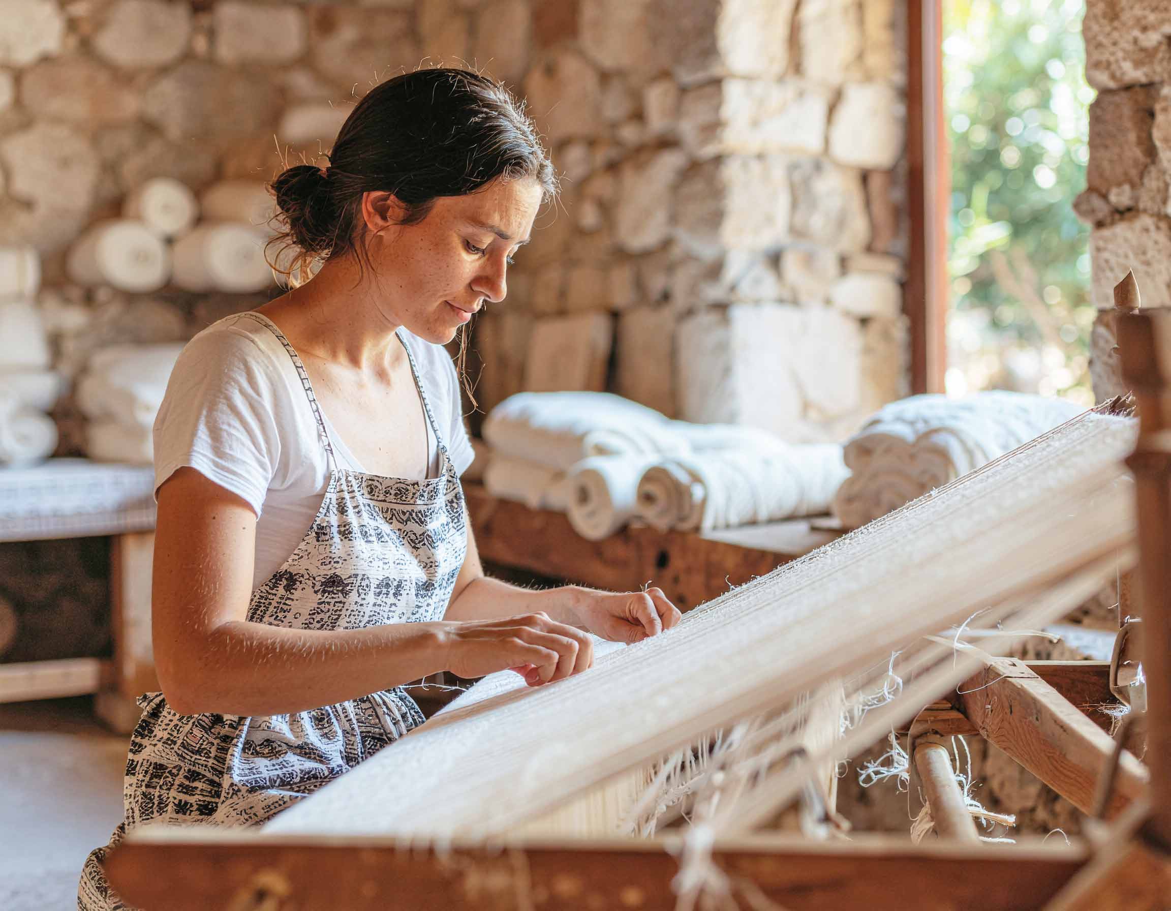 A woman weaving on a loom in a rustic setting with stone walls and natural elements. Sardinian artisan weaving traditional fabric on a loom for the sustainable fashion brand Antonello Tedde.