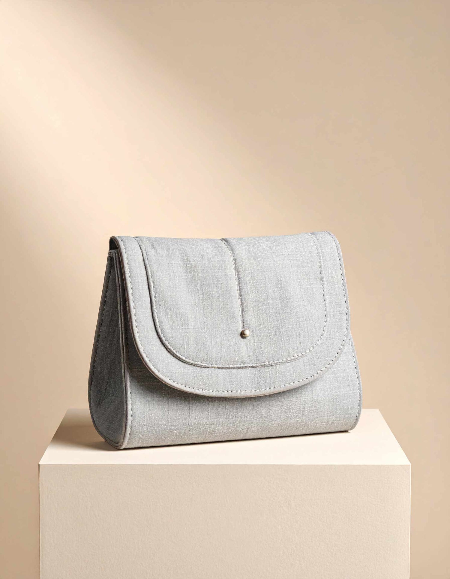 Light grey clutch bag on a beige pedestal with a beige background. Architectural mini handbag in matte neutral leather displayed on pedestal, representing 2026 sculptural handbag trend