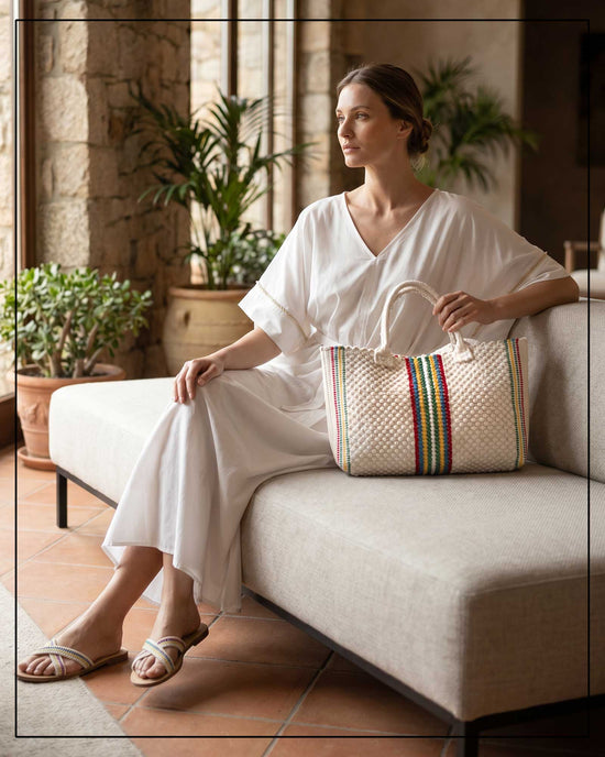 A woman is sitting on a couch holding an Antonello Tedde woven bag with colourful stripes in a cosy living room.